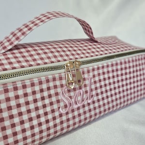 May include: A rectangular, pink and white gingham patterned cosmetic bag with a handle and a zipper closure. The bag has the embroidered name "Sel" in a cursive font. The zipper pulls are gold-colored.