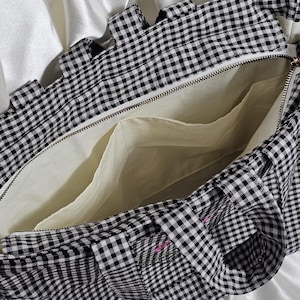 May include: Open view of a black and white gingham tote bag with a zipper closure. The interior is a light cream color with multiple pockets. The bag has a ruffled edge and is set against a white satin background.