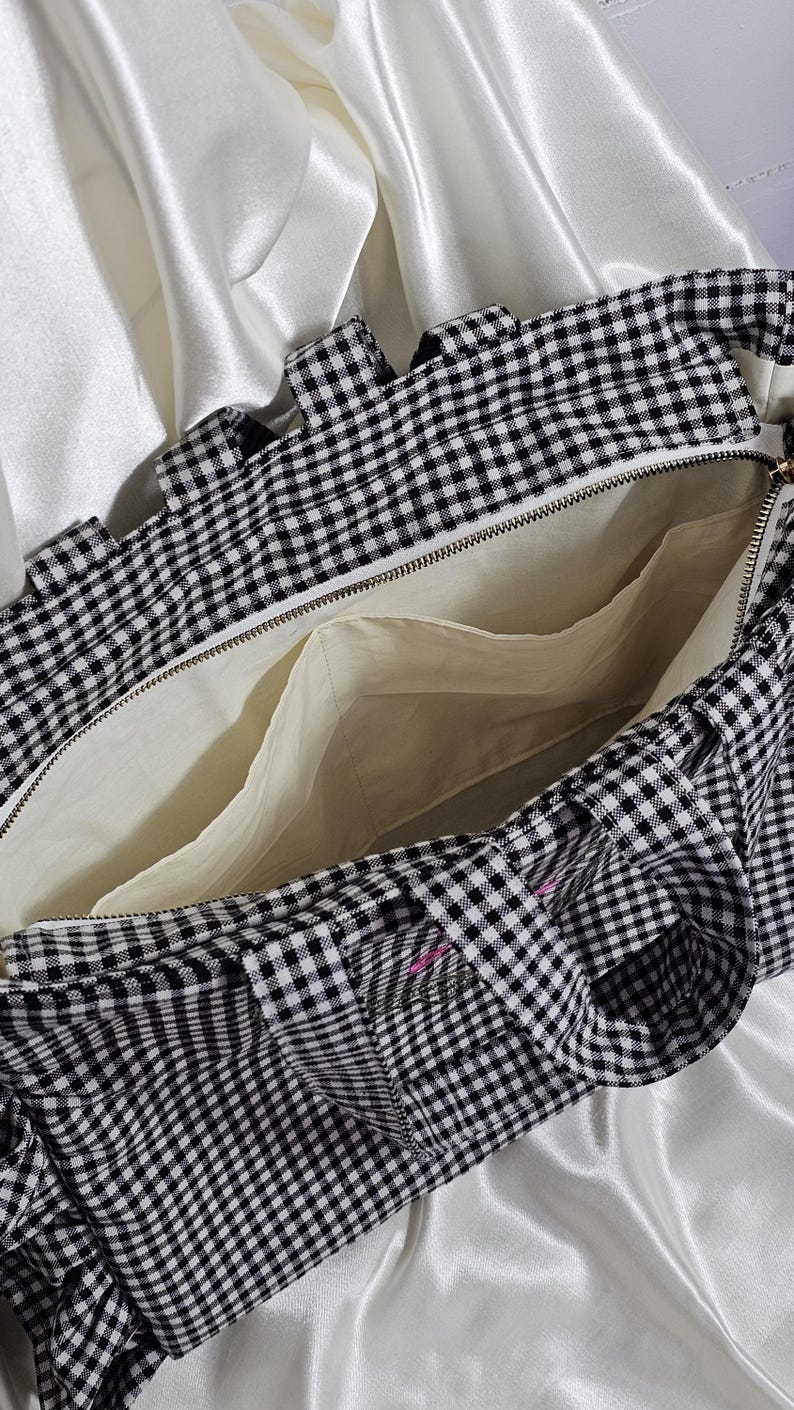 May include: Open view of a black and white gingham tote bag with a zipper closure. The interior is a light cream color with multiple pockets. The bag has a ruffled edge and is set against a white satin background.