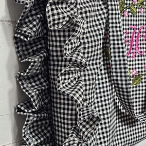 May include: Black and white gingham tote bag with a ruffled edge. The bag features a floral embroidery with the letter "M" in pink. The tote has handles for carrying.