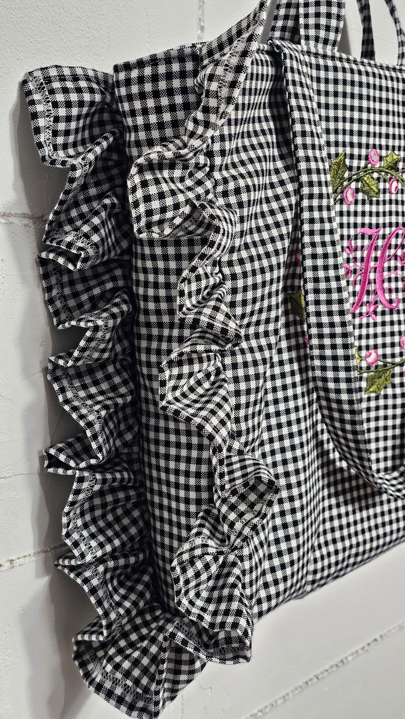 May include: Black and white gingham tote bag with a ruffled edge. The bag features a floral embroidery with the letter "M" in pink. The tote has handles for carrying.