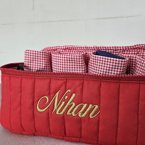 May include: Red quilted fabric organizer with a zippered top and gold embroidered text that reads "Nihan". The interior features red and white gingham compartments and a blue lining. A stylish storage solution.