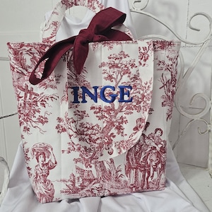 May include: A white tote bag featuring a red toile de jouy pattern of trees and figures, with the name INGE embroidered in blue thread on the front. The bag includes two fabric handles and a decorative dark red ribbon tied at the top center.