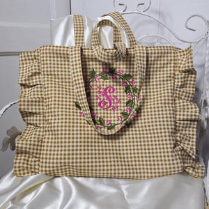 May include: A brown and white gingham tote bag with a floral wreath embroidery. The tote has a ruffle detail on the sides and a white satin lining. The wreath features pink flowers and a pink letter "S".