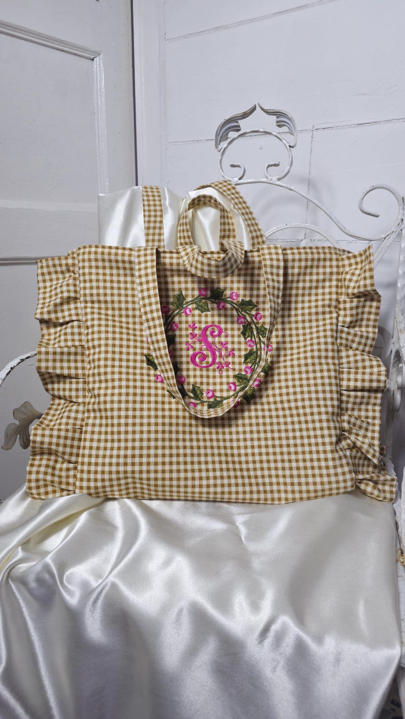 May include: A brown and white gingham tote bag with a floral wreath embroidery. The tote has a ruffle detail on the sides and a white satin lining. The wreath features pink flowers and a pink letter "S".