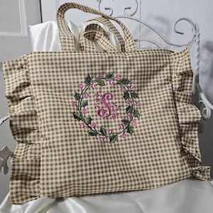 May include: A brown and white gingham tote bag with ruffled edges. The bag features a floral wreath embroidery with a pink letter "S" in the center. The tote has matching gingham handles.