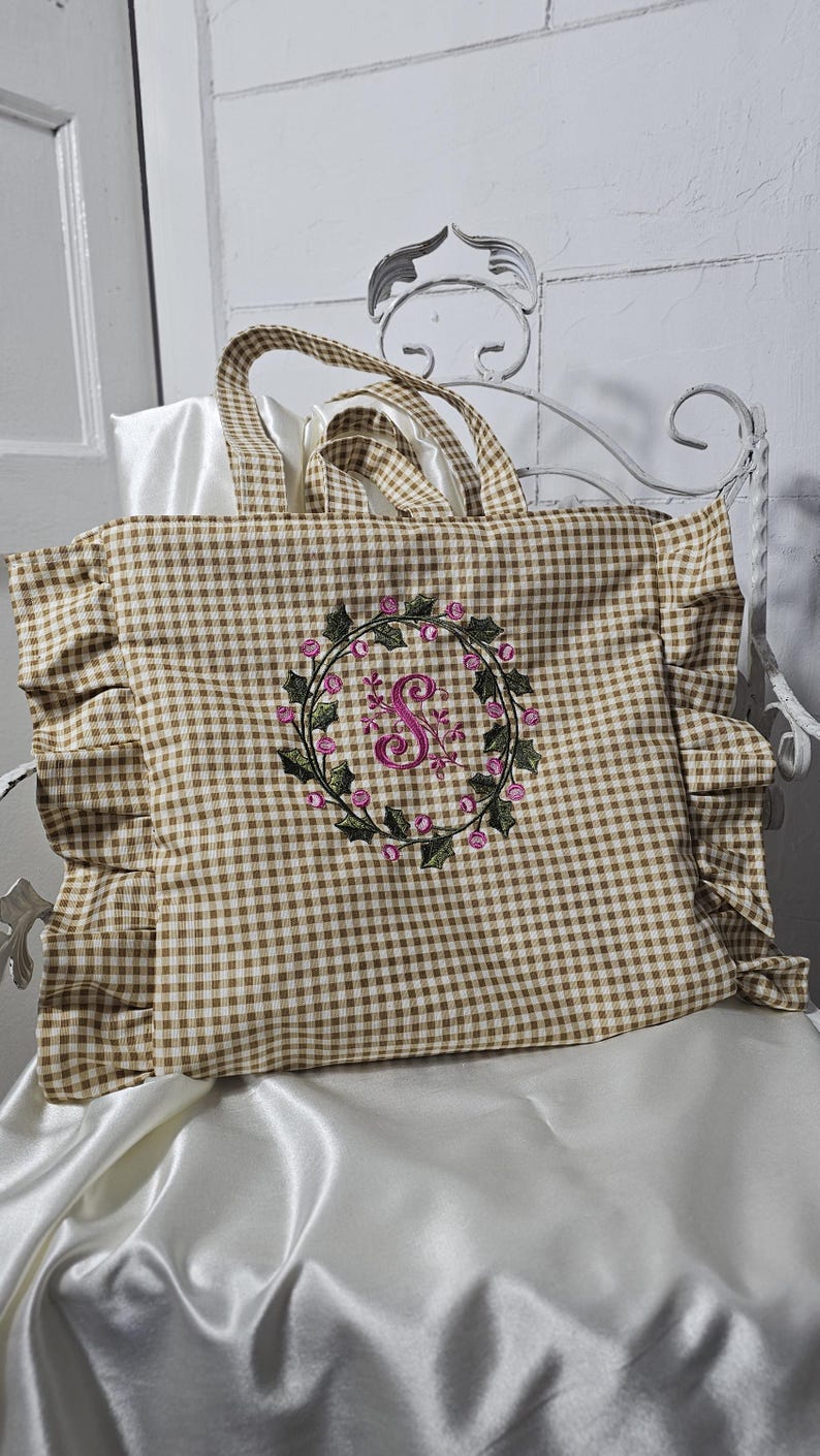 May include: A brown and white gingham tote bag with ruffled edges. The bag features a floral wreath embroidery with a pink letter "S" in the center. The tote has matching gingham handles.