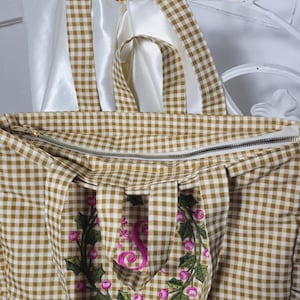 May include: A brown and white gingham tote bag with a zipper closure. The bag features a floral embroidery with the letter "S" in pink. The handles are made of the same gingham fabric and white satin ribbon.