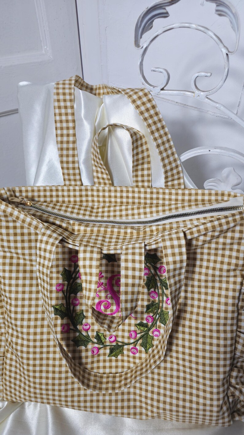 May include: A brown and white gingham tote bag with a zipper closure. The bag features a floral embroidery with the letter "S" in pink. The handles are made of the same gingham fabric and white satin ribbon.