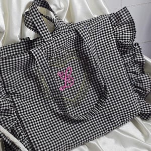 May include: Black and white gingham tote bag with a pink embroidered initial "L" inside a heart-shaped design. The tote features ruffled edges and sturdy handles for carrying. The bag is made of a soft fabric.