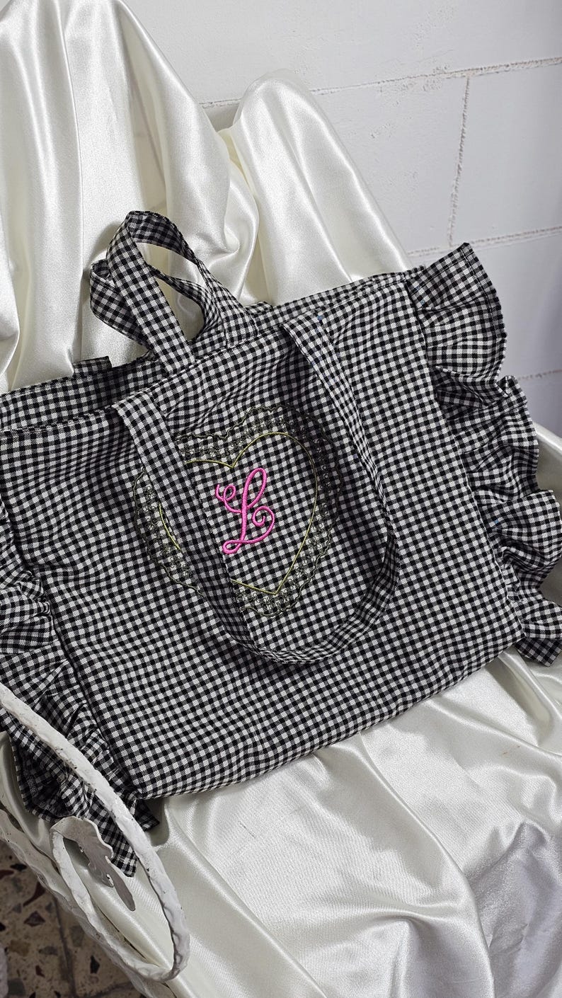 May include: Black and white gingham tote bag with a pink embroidered initial "L" inside a heart-shaped design. The tote features ruffled edges and sturdy handles for carrying. The bag is made of a soft fabric.