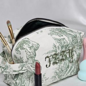 May include: A white toile makeup bag with a green pastoral print and the name Izzy embroidered in green script. The bag features a black zipper, two gold-handled makeup brushes, and a dark red lipstick standing in front of the pouch.