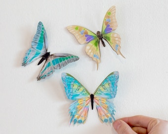 May include: Three paper butterflies with colorful wings. The butterflies are blue, green, yellow, and orange. They are attached to a white wall.