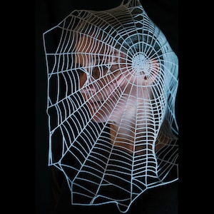 May include: A white, delicate spiderweb design, possibly a costume piece, is the focal point against a black background. The web is positioned in front of a person's face, creating a spooky, Halloween-themed effect. The web's intricate lines are clearly visible.