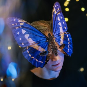 Puede incluir: Una persona con una máscara de mariposa azul y blanca. La máscara cubre los ojos y la nariz de la persona. La máscara está hecha de papel o cartón y tiene un diseño de mariposa realista.