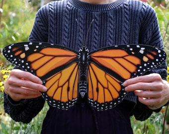 May include: A person is holding a large, orange and black monarch butterfly cutout. The butterfly has black spots and veins on its wings.