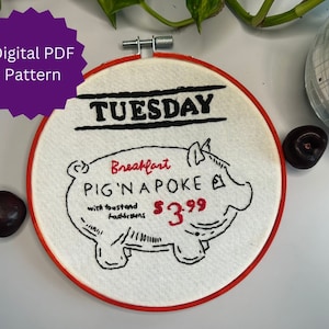 以下が含まれることがあります： 赤い縁取りのある刺繍の輪っかアート。豚の絵と「Breakfast Pig'n a Poke with toast and hashbrowns 3.99ドル」の文字、そして上部に「TUESDAY」の文字が書かれている。