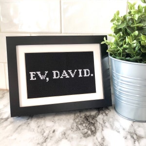 May include: A black framed picture with a white background and the text "EW, DAVID." in white letters. The picture is sitting on a white marble surface.