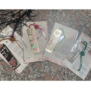 May include: Four Korean-style bookmarks in plastic packaging. The bookmarks have decorative designs and tassels in various colors. One bookmark has text in Korean and floral accents. The other bookmarks have intricate patterns.