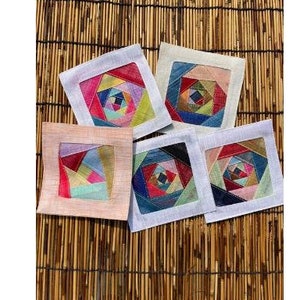以下が含まれることがあります： カラフルな布の正方形で幾何学模様が施された正方形の布製コースターが5つ。コースターは、編まれた竹の表面に一列に並べてあります。