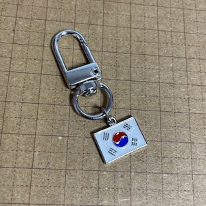 May include: A silver key chain with a South Korean flag charm. The flag is white with a red and blue yin-yang symbol in the center and three red bars on each side.
