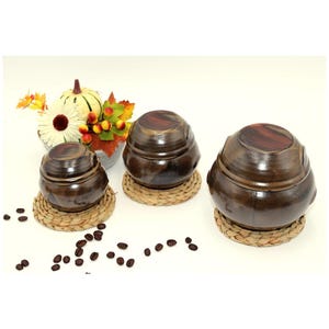May include: Three brown ceramic jars with wooden lids, each on a woven coaster. A small flower arrangement and scattered coffee beans are also present. The jars are of varying sizes.
