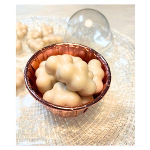 Puede incluir: Primer plano de un pequeño bol de color cobre lleno de malvaviscos cubiertos de chocolate blanco. El bol está sobre una superficie transparente y texturizada.