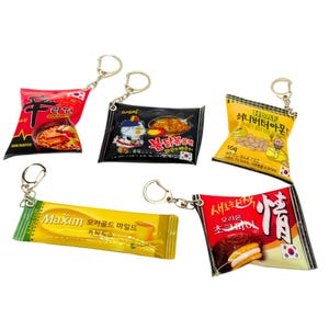 Pode incluir: Cinco chaveiros coloridos com lanches coreanos populares. Os chaveiros são feitos de plástico macio e flexível e apresentam imagens de macarrão instantâneo, mistura de café, amêndoas com manteiga de mel e biscoitos sanduíche de chocolate. Os chaveiros estão todos presos a argolas de chave prateadas.