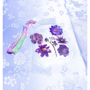 May include: A clear, mason jar-shaped pouch filled with pressed purple flowers. The pouch has a rainbow tassel attached to the top. The background is a light blue floral pattern.