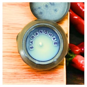 May include: A round, brown ceramic candle holder with a white candle inside. The candle holder has the word "SARANGHAE" spelled out in blue letters around the rim.