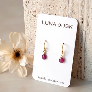 May include: Gold-coloured hoop earrings, each with a teardrop-shaped, deep pink gemstone. The earrings are presented on a white card with the text "LUNA DUSK" and the Etsy shop URL. A white flower is in the background.