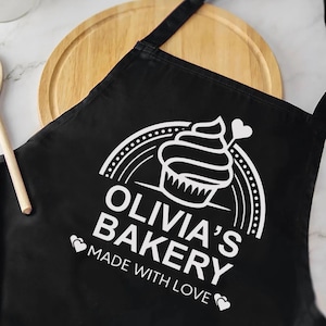 May include: Black apron with a white cupcake logo and the text "OLIVIA'S BAKERY MADE WITH LOVE". The apron is made of a durable fabric and is designed for use in a kitchen or bakery setting.