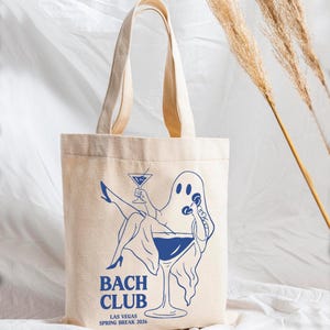 May include: A beige canvas tote bag featuring a blue graphic of a ghost sitting in a martini glass, holding a cocktail and a telephone. The text reads BACH CLUB LAS VEGAS SPRING BREAK 2026. The bag has two long shoulder straps and a rectangular body.