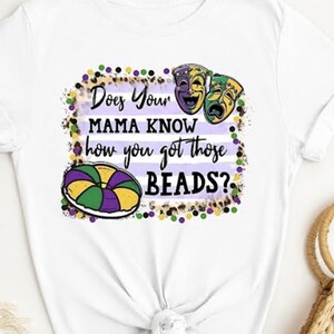 May include: White t-shirt with a purple, green, and gold Mardi Gras themed design. The design includes a king cake, beads, and two masks. The text on the shirt reads "Does Your Mama Know how you got those Beads?"