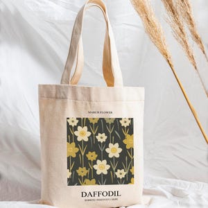 May include: A beige canvas tote bag featuring a floral design with daffodils in shades of yellow and white on a dark background. The text "MARCH FLOWER" is at the top, and "DAFFODIL" with "REBIRTH | POSITIVITY | HOPE" is at the bottom.
