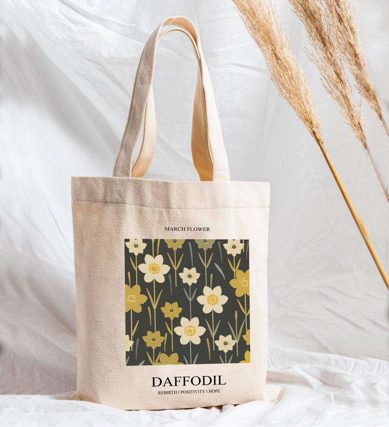 May include: A beige canvas tote bag featuring a floral design with daffodils in shades of yellow and white on a dark background. The text "MARCH FLOWER" is at the top, and "DAFFODIL" with "REBIRTH | POSITIVITY | HOPE" is at the bottom.