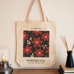 May include: A beige tote bag featuring a square design with red poinsettia flowers on a dark green background. The text "DECEMBER FLOWER" is at the top, and "POINSETTIA" with "CELEBRATION | GENEROSITY | UNITY" is at the bottom.