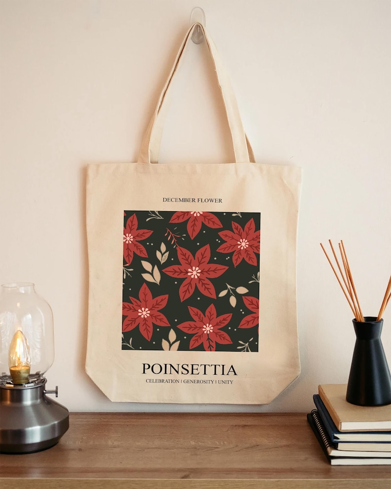 May include: A beige tote bag featuring a square design with red poinsettia flowers on a dark green background. The text "DECEMBER FLOWER" is at the top, and "POINSETTIA" with "CELEBRATION | GENEROSITY | UNITY" is at the bottom.