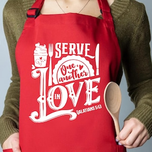 May include: A red apron with white text that reads "SERVE! One another IN LOVE Galatians 5:13". The design includes a cupcake, fork, knife, and wooden spoon. The apron is made of a durable fabric.