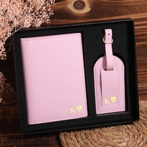 May include: A pink travel set including a passport holder and luggage tag, presented in a black gift box. Both items feature gold initials and a heart symbol. The set is displayed on a wooden surface with decorative elements.