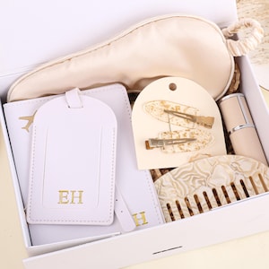 May include: A white gift box containing travel and beauty items. The contents include a cream-colored sleep mask, a luggage tag with gold initials, hair clips, a comb, and a small perfume bottle. The items are in cream and gold tones.