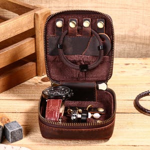 May include: Open, brown leather travel jewelry case with a zipper. Inside are a watch, rings, earrings, and bracelets. The case is on a wooden surface.