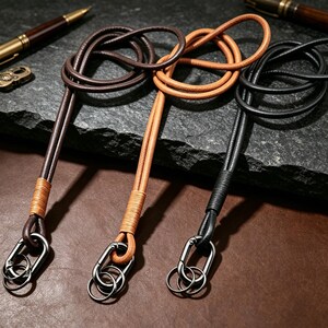 Puede incluir: Tres cordones de cuero en marr&oacute;n oscuro, tostado y negro. Cada cord&oacute;n presenta un acabado de cuero enrollado cerca de la base, un mosquet&oacute;n de metal oscuro y una anilla met&aacute;lica circular en el extremo para sujetar llaves u objetos.