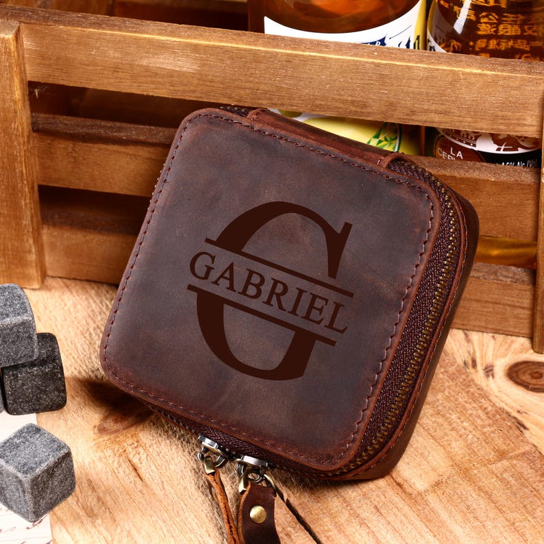 May include: A brown leather square case with a zipper closure. The case is embossed with the name "GABRIEL" and a large letter "G". The case is sitting on a wooden surface. Beside the case are several gray stone cubes.