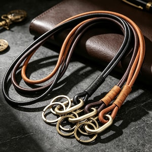 May include: Three leather lanyards in black, dark brown, and tan, each featuring a wrapped cord section and a matte gold-toned metal carabiner attached to a circular split ring. The cords are smooth and rounded, designed for holding keys or small items.