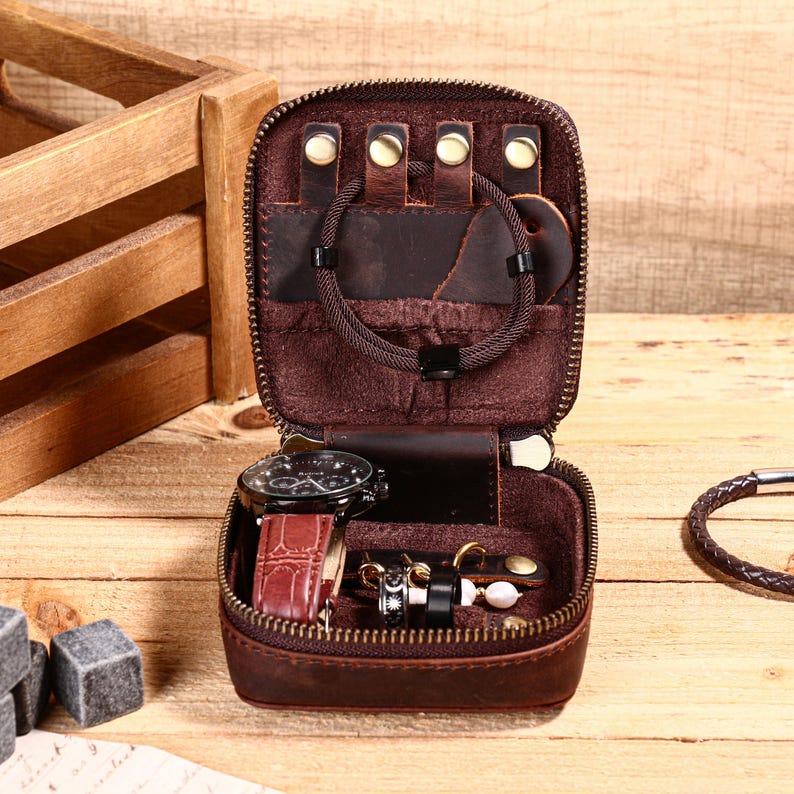 May include: Open, brown leather travel jewelry case with a zipper. Inside are a watch, rings, earrings, and bracelets. The case is on a wooden surface.