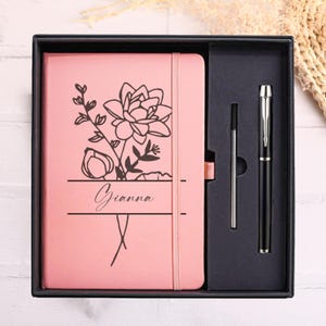 Puede incluir: Un juego de cuaderno y bolígrafo rosa en una caja de regalo negra. El cuaderno presenta un diseño floral y el nombre "Gianna". El bolígrafo es negro con detalles plateados. Incluye un recambio.