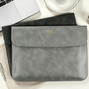 May include: A rectangular gray laptop sleeve with a flap closure and the gold embossed letters HA centered on the front. The sleeve features a smooth, matte finish and is positioned over a black laptop sleeve of the same design and size.