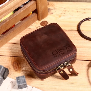 May include: A brown leather square zippered case with the name "GIBSON" and the text "BEST DAD EVER" engraved on the front. The case has a bronze-colored zipper and pull tabs. The background includes a wooden crate and a braided bracelet.