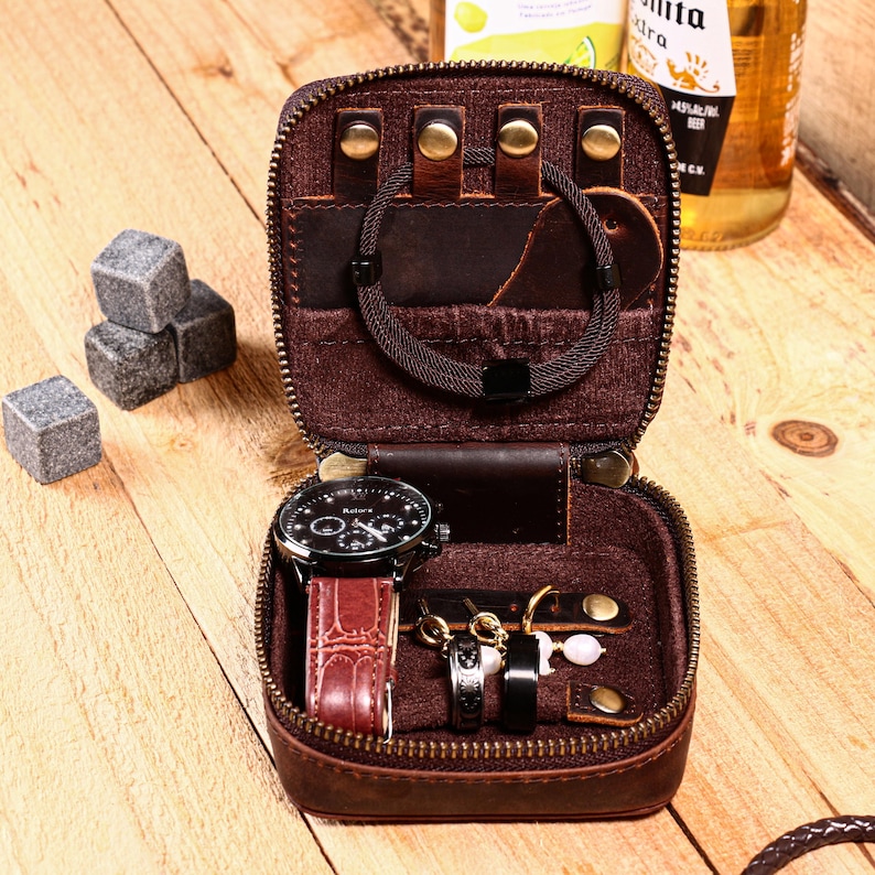 May include: A brown leather travel jewelry case with a zipper, holding a black watch with a red band, rings, earrings, and a bracelet. Gray stone cubes are next to the case. A bottle of beer is in the background.